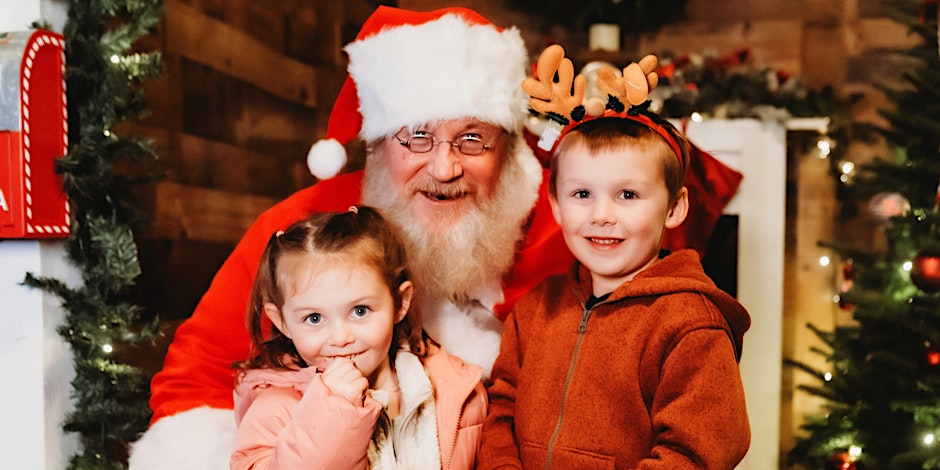 Santa Claus smiling at camera with two young kids on his lap.